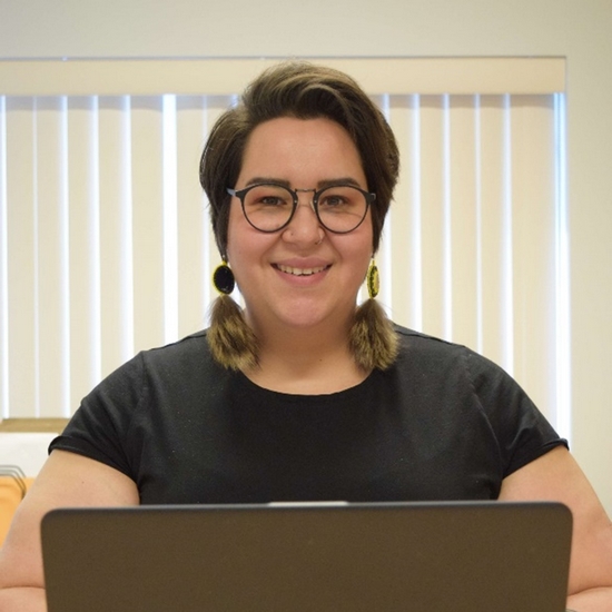 Alexia smiling at a laptop. She has short, dark hair, glasses, and fur earrings.
